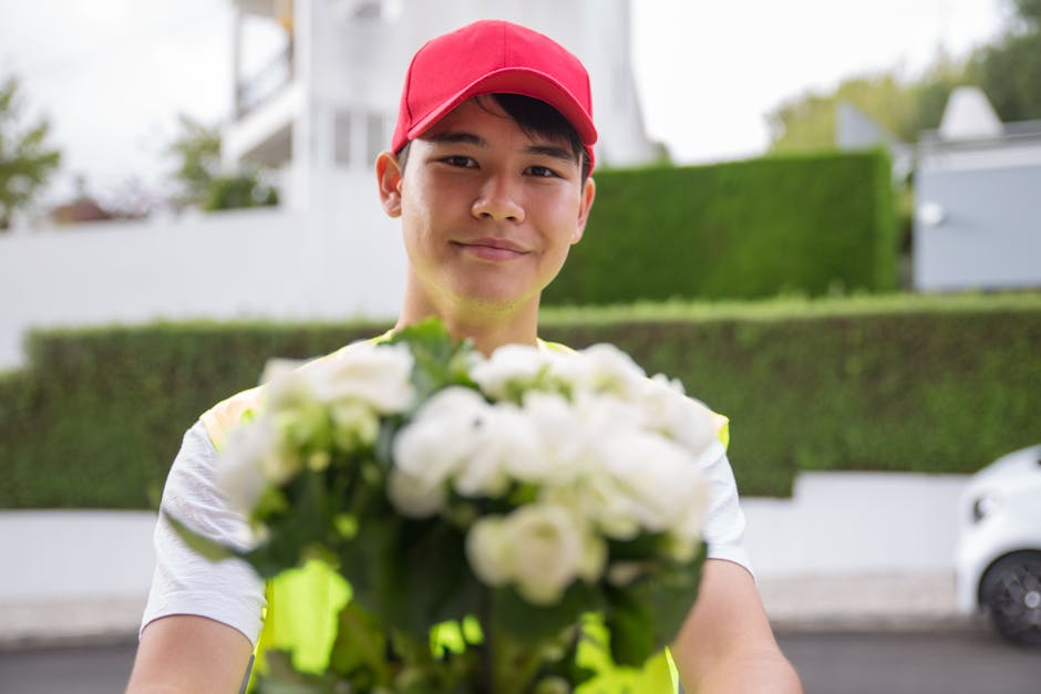 A delivery person wearing a red cap and a yellow high-visibility vest holds a bouquet of fresh white flowers in front of them, with their face smiling gently. The bouquet features white roses, lisiant