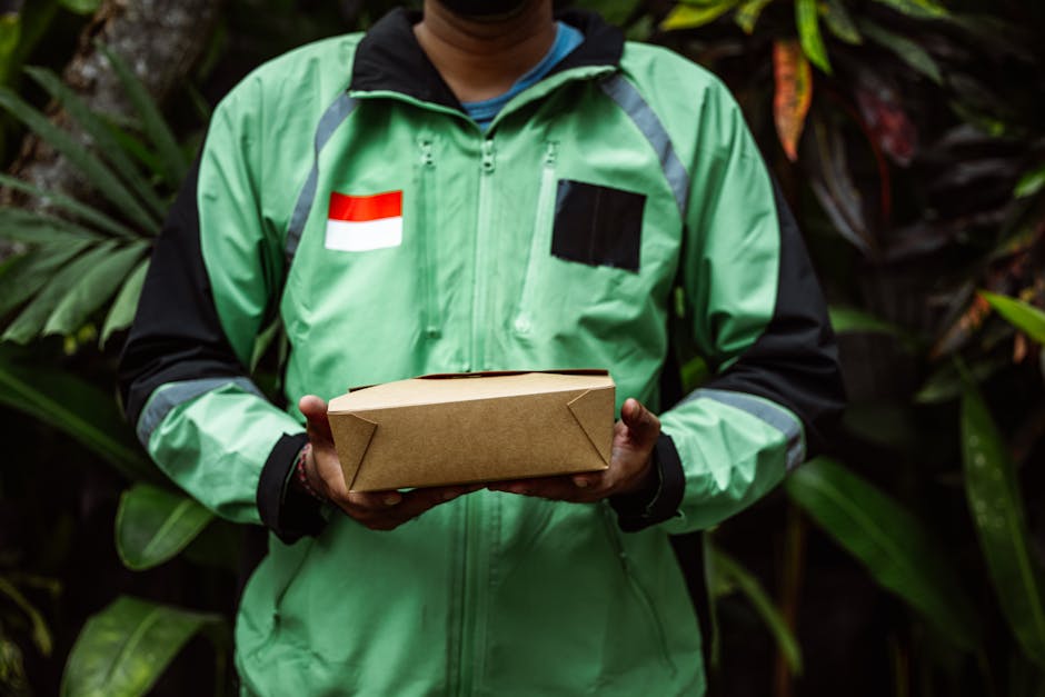 A delivery person dressed in a bright green jacket with a Dutch flag patch on the chest is holding a neatly wrapped rectangular parcel in their hands. The person is standing outdoors amid lush, green 