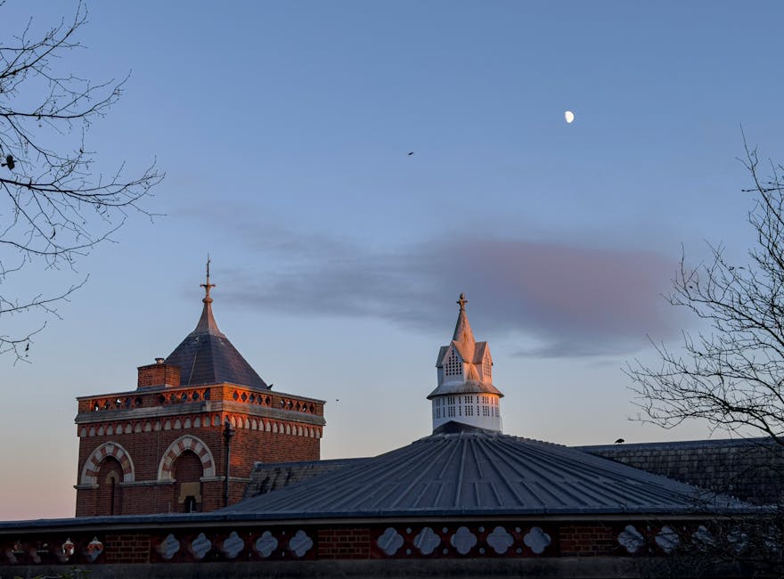 A view of historic brick and white wooden church towers against a twilight sky, with the full moon visible overhead. Bare tree branches frame the scene, and soft evening light casts a warm glow on the