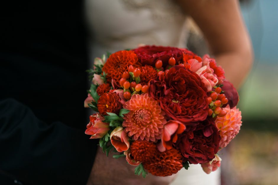 A vibrant floral bouquet featuring a mix of deep red and coral-colored flowers, including large peonies, dahlias, and carnations, accented with small orange berries and green foliage. The flowers are 