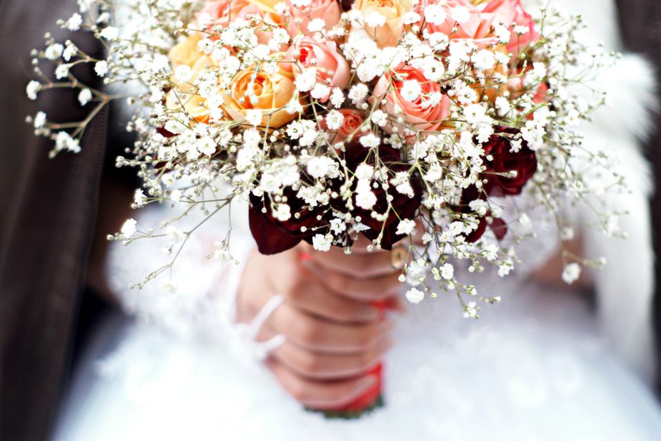 A close-up of a bridal bouquet featuring peach and pink roses, surrounded by delicate white baby's breath flowers and dark burgundy foliage. The bouquet is held by a bride dressed in white, with a gli