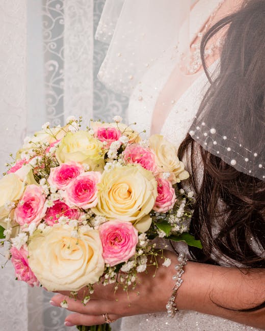 A bride in a white lace wedding dress and veil is holding a floral bouquet composed of cream and pink roses, with sprigs of baby's breath and green foliage. The bouquet features large, fully bloomed r