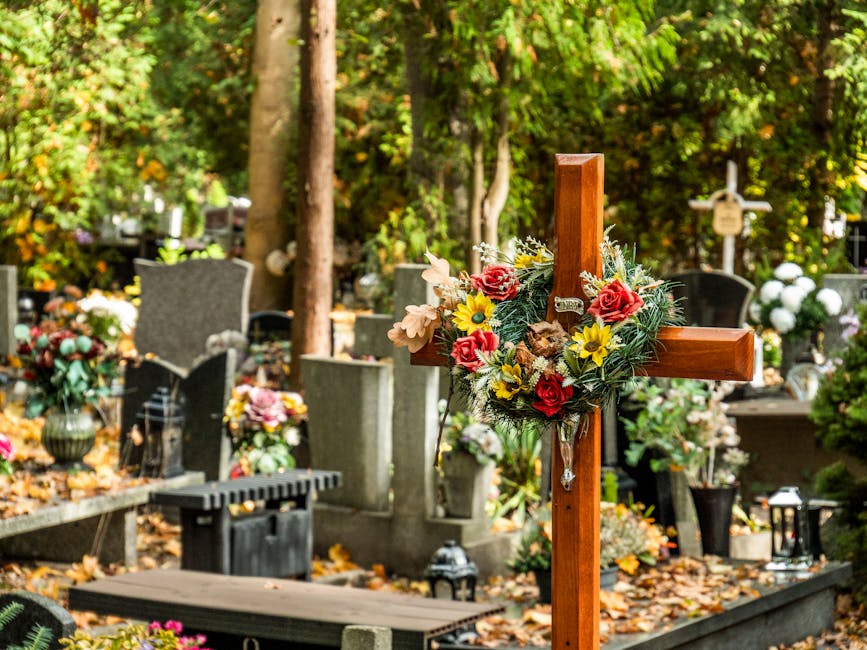 A wooden funeral cross adorned with a floral arrangement featuring red roses, yellow daisies, pale pink lilies, and green foliage. The cross is positioned upright in a cemetery setting with other grav
