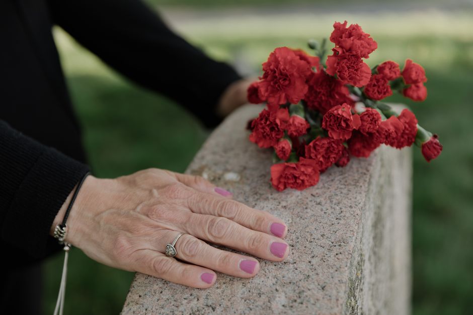A close-up view of a woman's hand resting on a stone memorial, adorned with a silver ring and a black bracelet. Next to her hand, placed on the memorial, is a small bouquet of red carnations with ruff