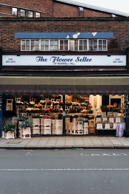 The storefront of The Flower Seller of Muswell Hill, displaying a vibrant floral arrangement with a variety of fresh flowers in shades of yellow, orange, pink, and purple arranged on white shelves and