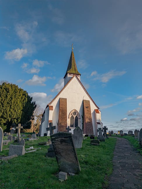 A small white church with a steep, dark roof and a tall, pointed spire topped with a weather vane, situated among green grassy grounds and numerous weathered gray gravestones leaning at various angles