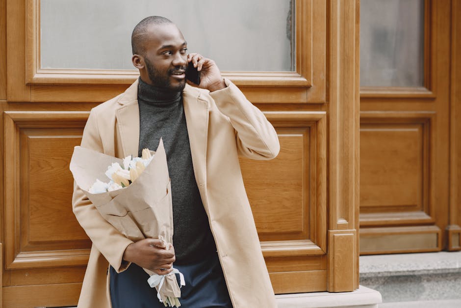A man with dark skin, short hair, and a beard is standing outdoors against a wooden-paneled wall, holding a bouquet of fresh, white flowers wrapped in beige paper. He is dressed in a beige overcoat ov